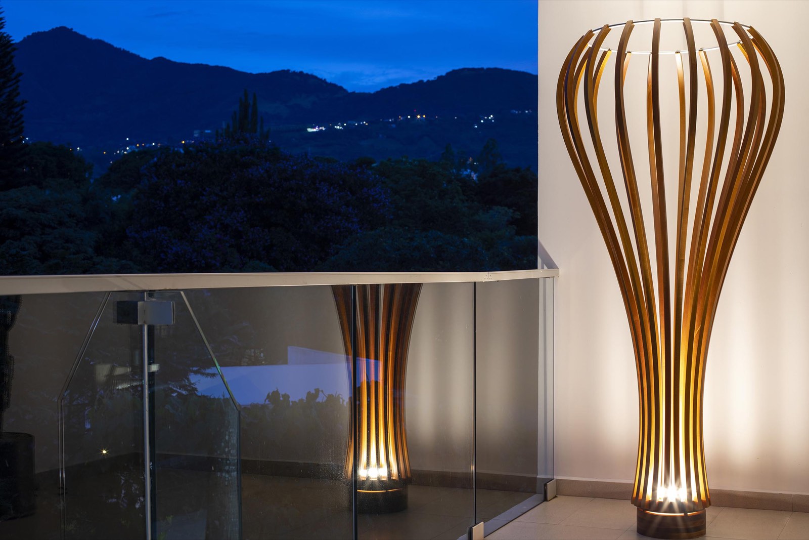 Glow of Grace sculptural cedar floor lamp glowing warm on a terrace at dusk, mountains behind